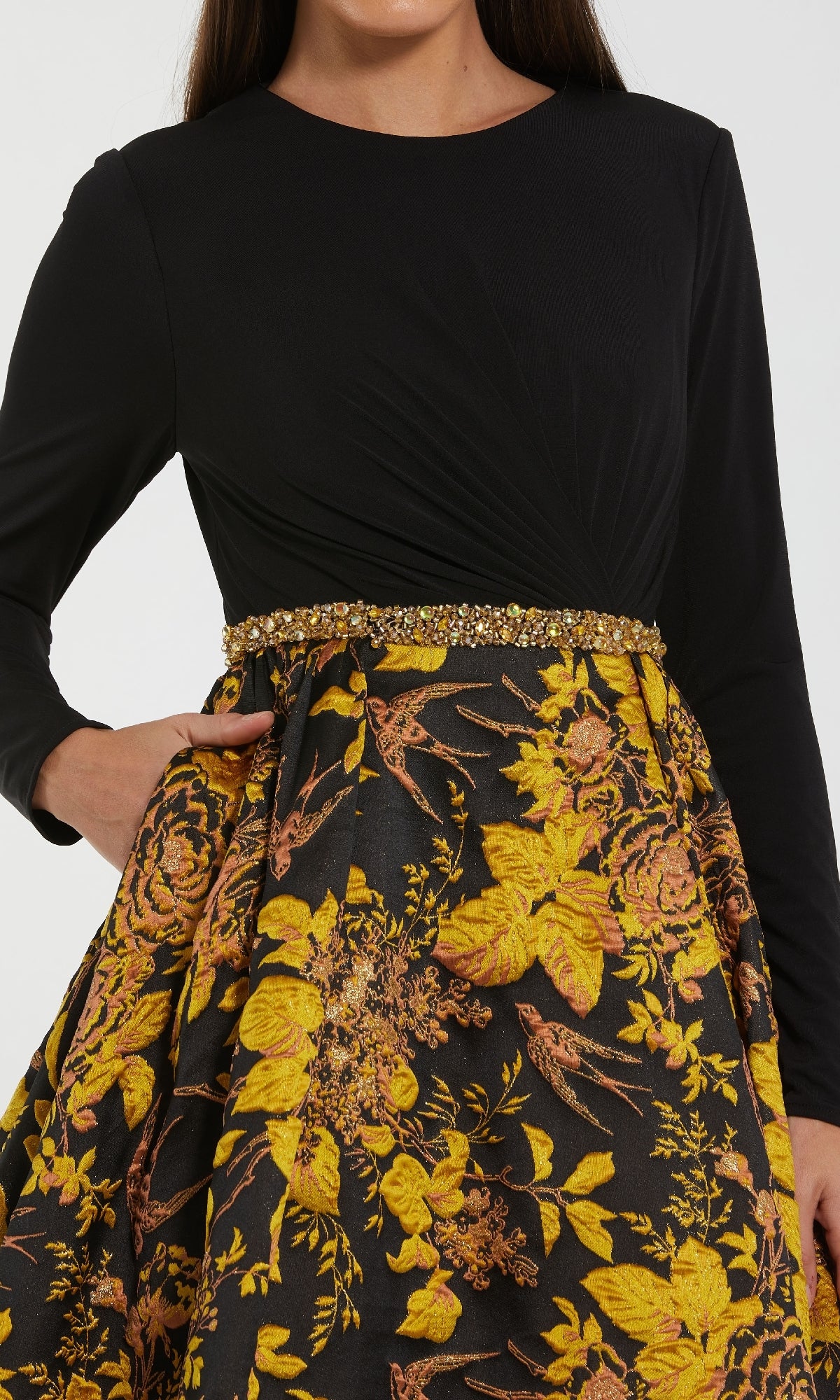 A woman wears the Black and Gold Formal Dress: Mac Duggal 69000, featuring a modest black long-sleeve top and a high-waisted gold floral skirt with a beaded waistband. Her right hand rests casually in her pocket, exuding elegance.